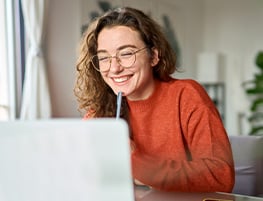 Young woman in glasses and an orange sweater smiling at a laptop screen