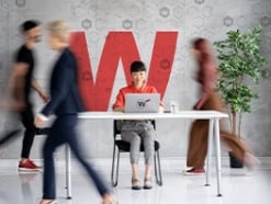 Woman working on her laptop at a white table with blurred people walking around her